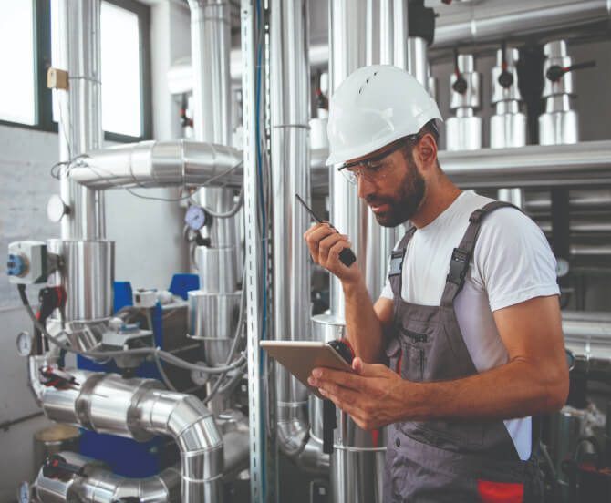 Engineer holds tablet and walkie-talkie in boiler room.