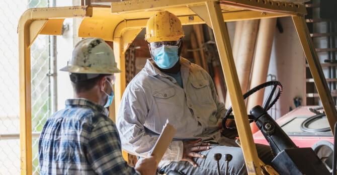 Two male warehouse workers wearing protective face masks talking. One is seated on a forklift.