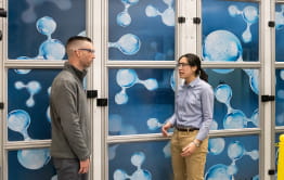 Two Enbridge Gas employees in safety goggles standing in front of large graphics of hydrogen molecules.