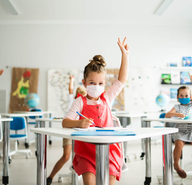Young children with protective face masks on sitting at desks in a classroom, raising hands