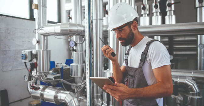 Engineer holds tablet and walkie-talkie in boiler room.