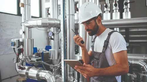Engineer holds tablet and walkie-talkie in boiler room.