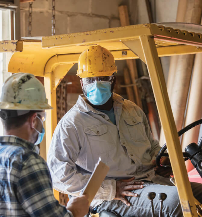 Two male warehouse workers wearing protective face masks talking. One is seated on a forklift.