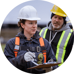 Two HVAC professionals in hard hats and safety vests have a discussion. One holds a clipboard, while the other looks on.