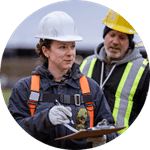 Two HVAC professionals in hard hats and safety vests have a discussion. One holds a clipboard, while the other looks on.