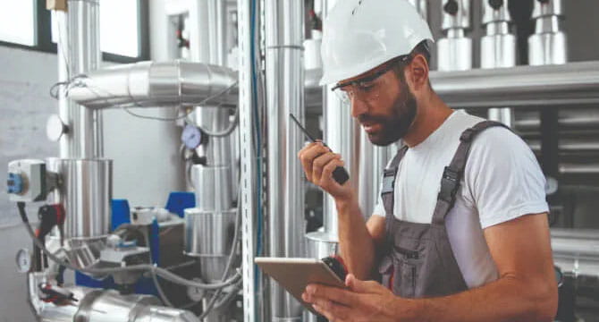 Engineer holds tablet and walkie-talkie in boiler room.