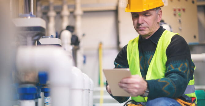 Man in hard hat kneeling in boiler room holding tablet