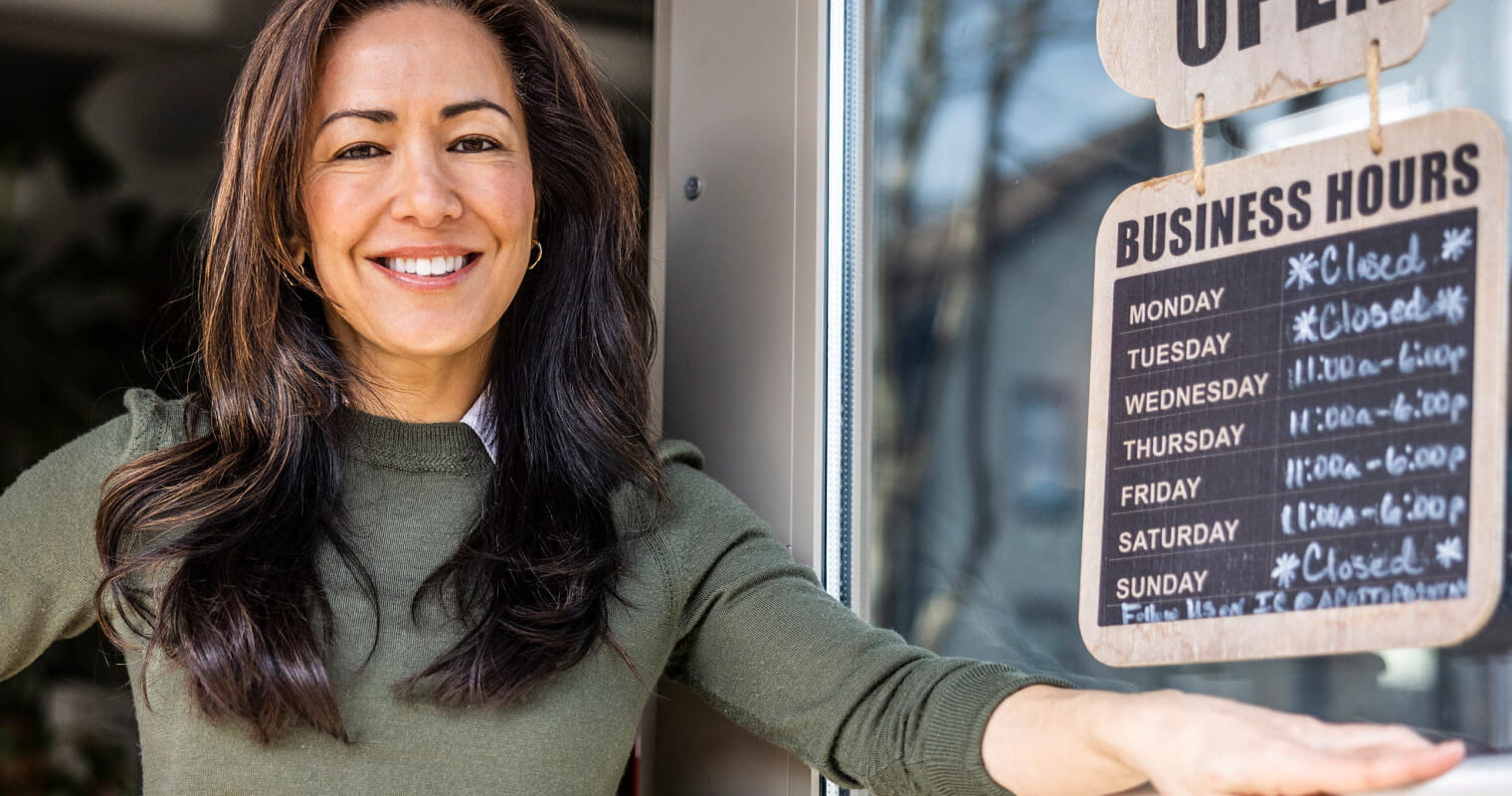 A shop owner stands in the doorway of their establishment with the door ajar. On the door, an open sign displaying business hangs on the back.