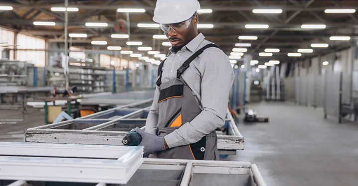 A worker in a hard hat and protective overalls works with metal frames in a large industrial warehouse. The setting is spacious, with overhead lights and rows of machinery and materials in the background.