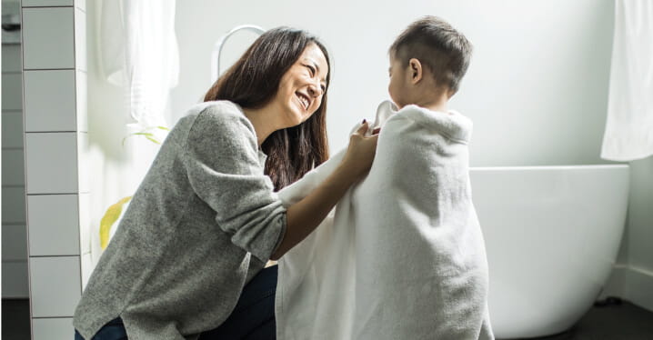 A parent kneels in a modern bathroom and smiles as she wraps her young child in a white towel. The child stands near a freestanding bathtub, and the setting is clean, cozy and softly lit.