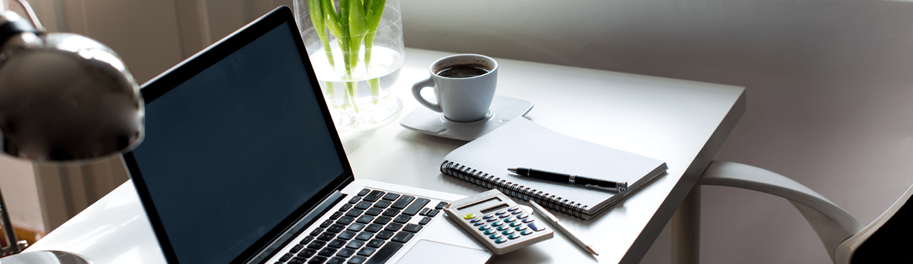 A desk with laptop with a calculator, pen, diary and a cup of coffee on it.